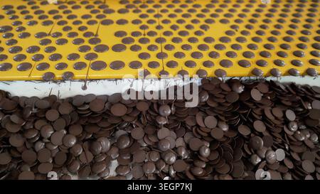 Industrial process making chocolate drops. Liquid cocoa drips onto yellow conveyor forming dots. Finished brown candy chips collect below in pile. Foo Stock Photo