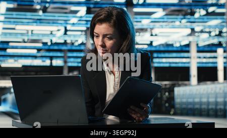 Data center software developer optimizing equipment rigs using devices. Woman in server farm running programming script on laptop and tablet, adjusting system parameters Stock Photo