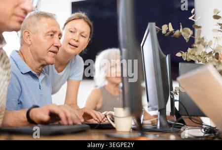 Young female teacher of computer class for seniors assisting man Stock Photo