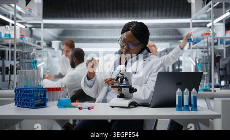 Clinical laboratory technologist pipetting reactant into blue solution for purity analysis. African american woman adding liquid agent to test tube to observe chemical reaction properties, camera A Stock Photo