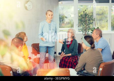 Older woman presides over support group meeting with mature people listening Stock Photo