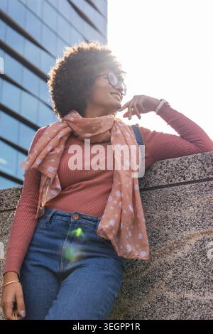 African American woman leaning on concrete railing in city plaza looking up wearing polka-dot scarf Stock Photo