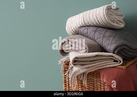 Wicker basket with stack of different cozy blankets on turquoise background Stock Photo