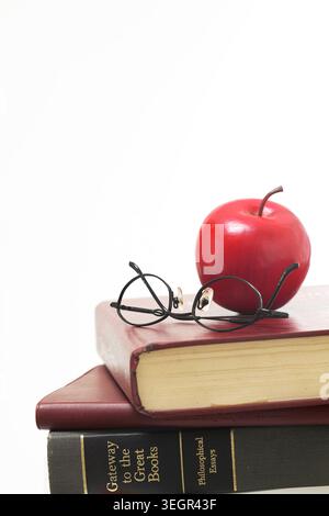 A pair of reading glasses lies on top of a stack of books, with a red apple and a pair of glasses resting on top. Stock Photo