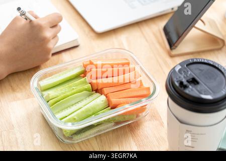 A glass container with a mix of sliced green and orange vegetables on a wooden desk, surrounded by a laptop, a hand holding a pen, and a coffee cup. Stock Photo