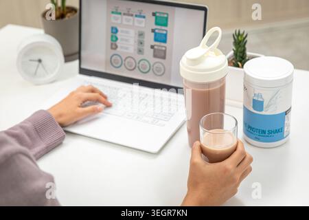 A person sits at a desk, holding a glass of protein shake while working on a laptop. Stock Photo