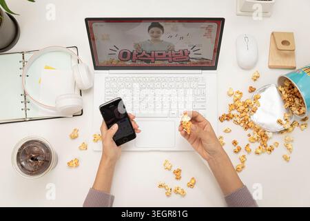 A person sits at a desk, working on a laptop, surrounded by popcorn, while watching a video on the screen. Stock Photo