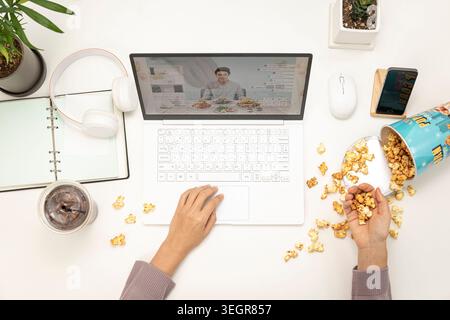 A person is using a laptop on a white desk, surrounded by various items such as a plant, phone, and popcorn. Stock Photo