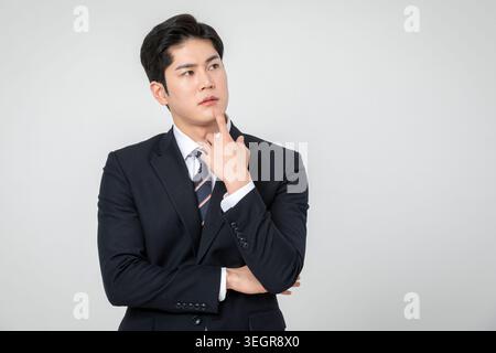 A young Asian businessman in a suit looks thoughtful, touching his chin, against a neutral background. Stock Photo