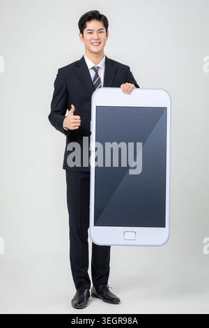 A young Asian businessman in a suit is holding up a large smartphone with a blank screen, giving a thumbs up. Stock Photo