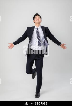 A young Asian business executive is shown in a triumphant pose, arms outstretched and eyes closed, in a modern office setting. Stock Photo