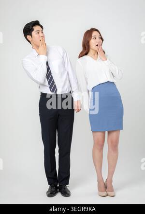 A young Asian man and woman stand together, both covering their mouths with their hands, looking surprised. Stock Photo