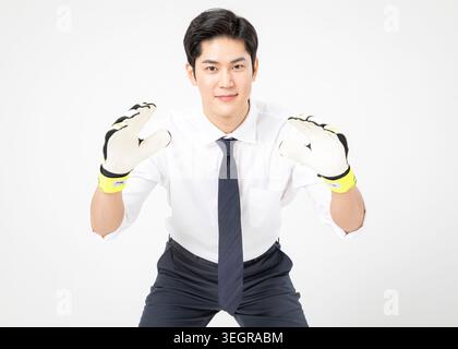 A young Asian man in a white shirt, black tie, and dark pants, wearing goal-keeper gloves, poses in front of a plain white background. Stock Photo