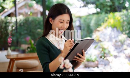 young Asian women writing notes on digital tablet at outdoor cafe garden terrace for business planning study research Stock Photo