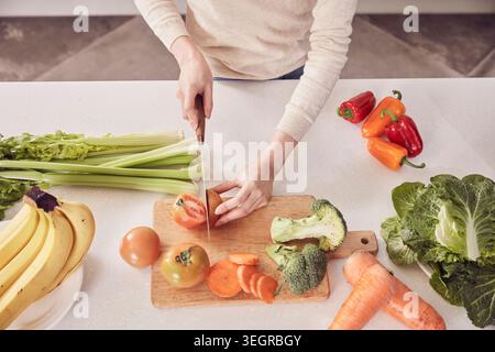 A woman is chopping tomatoes on a kitchen counter surrounded by various vegetables and fruits. Stock Photo