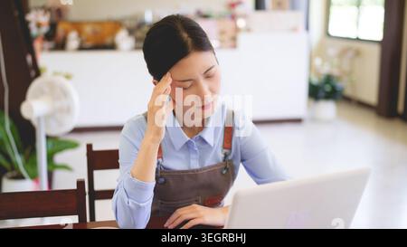 Young Asian women cafe owner feeling stress headache while using laptop managing small business restaurant workspace Stock Photo