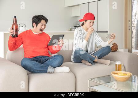 A man and a woman are sitting on a couch, watching a game and interacting with their devices while enjoying snacks and drinks. Stock Photo