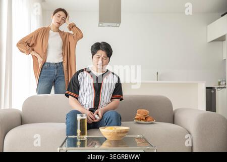 A young man sits on a couch, playing a video game, while his girlfriend stands behind him, looking bored and uninterested. Stock Photo