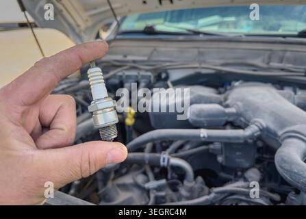 Close up of a mechanic hand holding a used spark plug near an open car engine bay during vehicle maintenance and ignition system inspection. Stock Photo