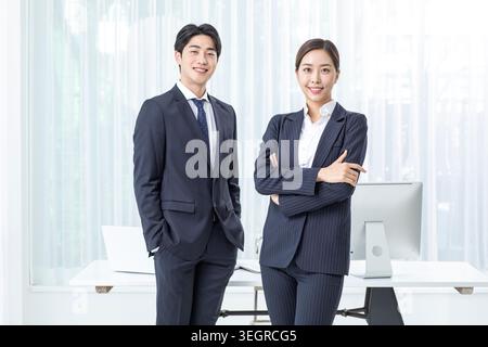 Two young, professional colleagues in business attire stand smiling in a modern, well-lit office. Stock Photo