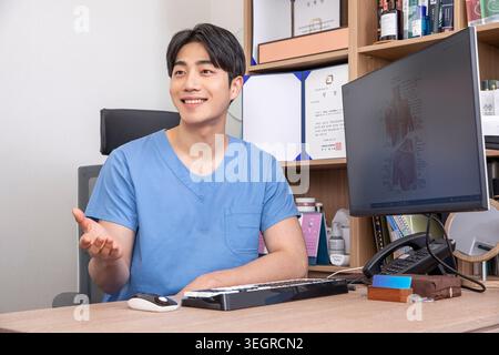A young Asian stock image editor sits at his desk, smiling as he works on a computer in a modern, well-lit office. Stock Photo
