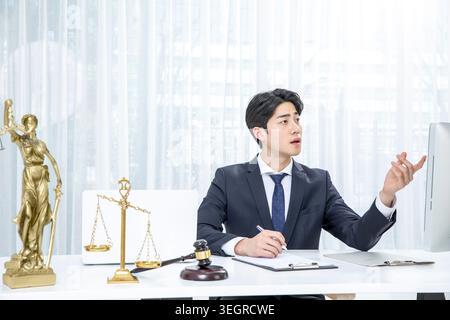 A young Asian lawyer sits at a desk in a modern office, studying a document with a serious expression. Stock Photo