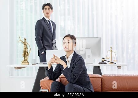 A young Asian woman in a business suit sits on a couch in a modern office, looking at a screen with a serious expression. Stock Photo