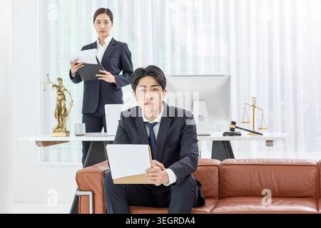 A young Asian lawyer sits on a couch in a modern law office, holding a folder and looking at a tablet while a female colleague stands behind him, reviewing documents. Stock Photo