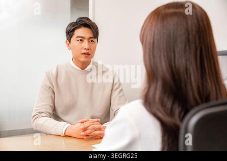 A young Asian man and woman sit across from each other at a wooden table, their hands clasped in a calm, professional meeting. Stock Photo