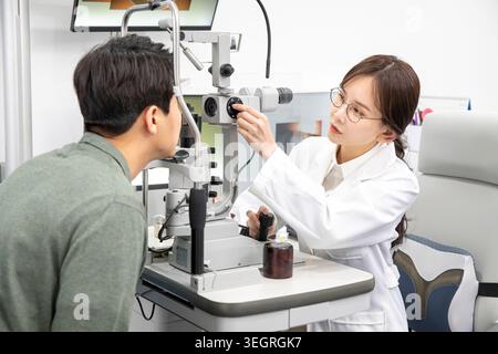 A young Asian woman, wearing a white lab coat, examines a patient's eyes with an ophthalmic instrument in a modern clinic. Stock Photo
