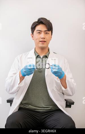 A young Asian man in a white lab coat and gloves holds a pair of glasses in a sterile laboratory setting. Stock Photo