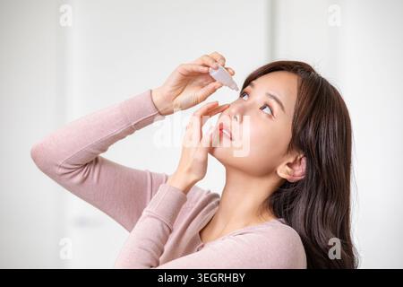 A young woman is carefully applying eye drops to her right eye in a clean and well-lit bathroom. Stock Photo