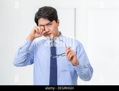 A young Asian man in a white shirt and blue tie rubs his tired eyes in a modern office setting. Stock Photo