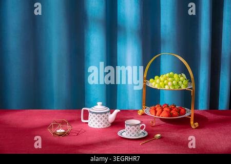 A delicate tea set, a gold two-tiered serving stand, and a selection of fresh fruit are arranged on a rich, red velvet tablecloth in a warm, sunlit room. Stock Photo