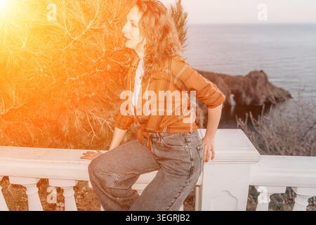 Woman ocean sunset young lady in plaid shirt sitting on white stone balcony overlooking calm sea during golden hour Stock Photo