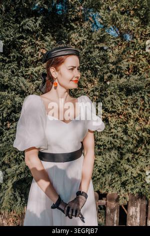 Woman vintage dress garden posing gracefully in a white puff sleeve gown with black accessories against lush green foliage during golden hour Stock Photo