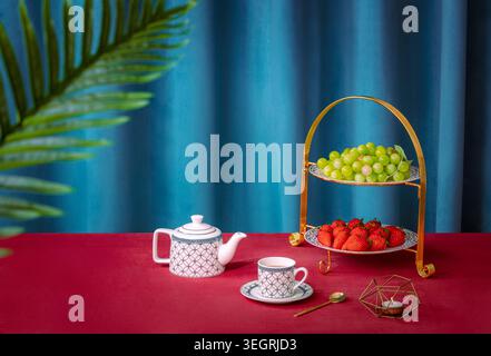 A luxurious gold two-tier serving stand holds a selection of fresh fruit and fine china tea cups on a velvet table in a luxurious setting. Stock Photo