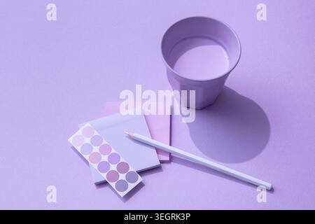 A cup of water, a white pencil, and a set of colored sticky notes are arranged on a purple background. Stock Photo