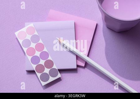 A white pencil lies on a stack of pastel pink and purple paper, next to a cup of water and a sheet of circular stickers. Stock Photo
