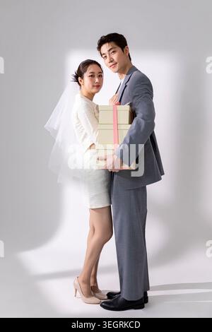 A young couple in formal attire, holding a gift, posing together in a neutral setting. Stock Photo