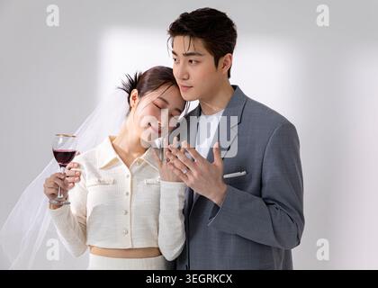 A young Asian couple in a wedding outfit share a romantic moment, holding wine glasses and gazing at each other. Stock Photo