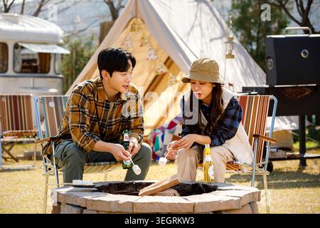 A young couple is roasting marshmallows over a campfire in a sunny, rustic outdoor setting. Stock Photo