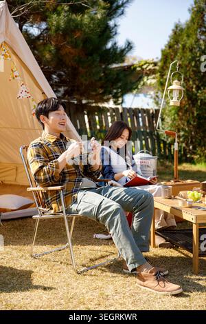 A young couple relaxes in a campsite, enjoying coffee and reading books in the warm sunlight. Stock Photo