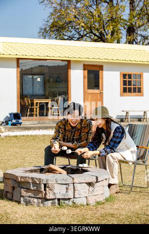 Two friends are roasting marshmallows over a campfire in a sunny backyard. Stock Photo