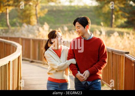 A young woman and man walk hand in hand on a wooden bridge in a sunny park. Stock Photo
