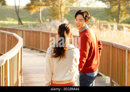 A young couple walks hand in hand on a wooden bridge in a sunny forest. Stock Photo