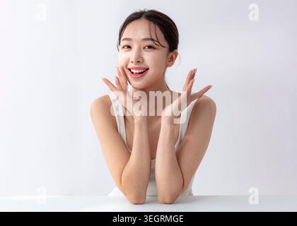 A young Asian woman sits at a white table, smiling and touching her face with both hands. Stock Photo
