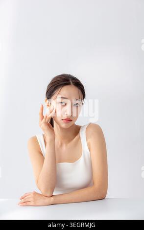 A young Asian woman sits in a white studio, gently touching her face with her right hand. Stock Photo