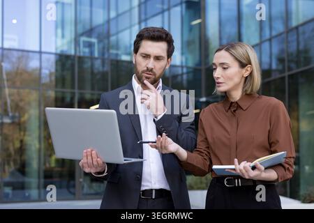 Business colleagues collaborating on an important project, analyzing data on a laptop, and brainstorming solutions while standing outside a modern off Stock Photo