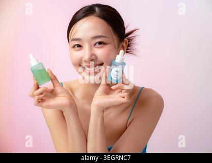 A young Asian woman is smiling and holding two skincare bottles against a soft pink background. Stock Photo